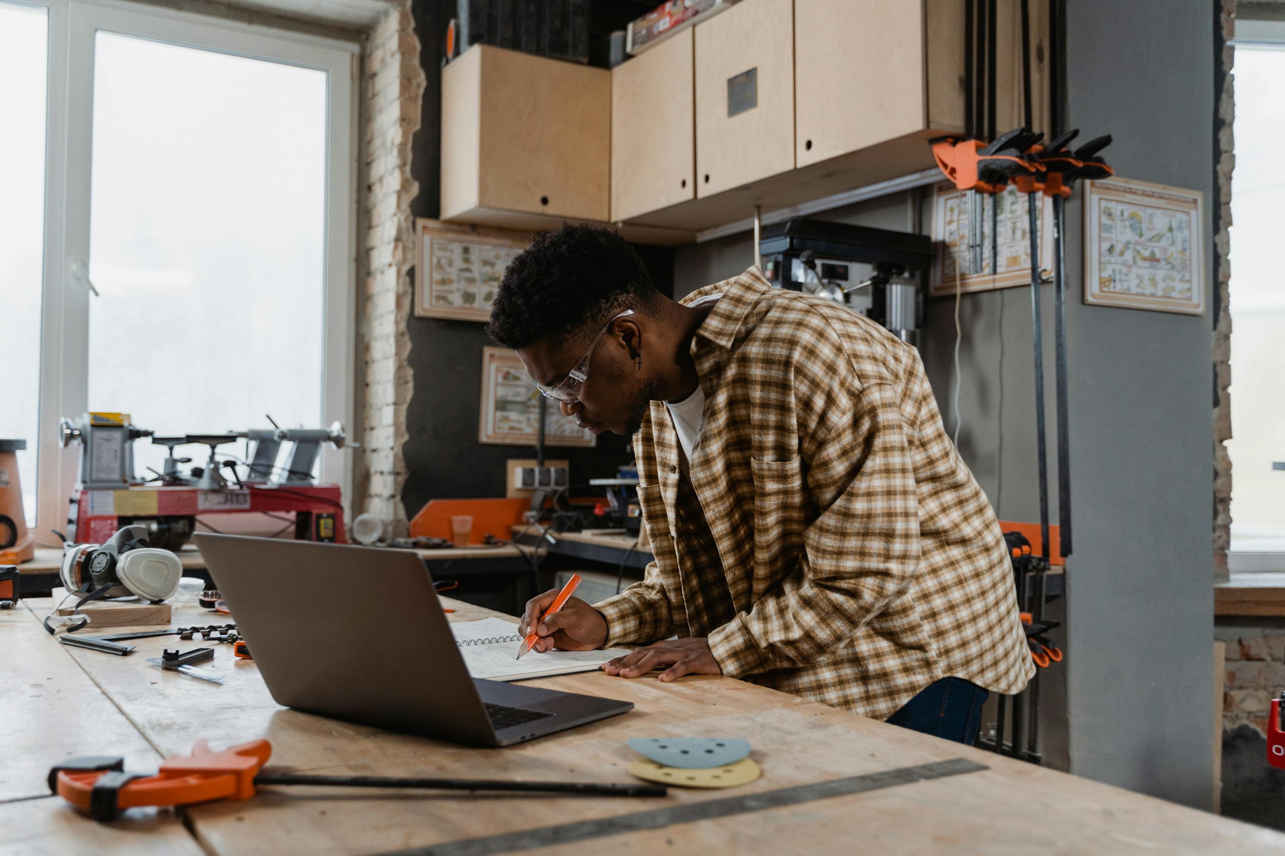 Carpenter in a workshop drafting plans on a laptop surrounded by tools and woodworking equipment.